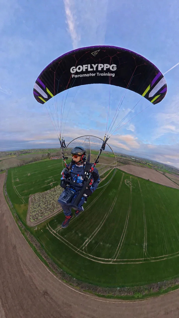 Paramotor pilot flying over green fields, paragliding wing with visible printed vinyl GOFLYPPG graphic.
